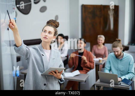 Waist up portrait of contemporary young woman pointing at whiteboard while teaching training course for students in class, copy space Stock Photo