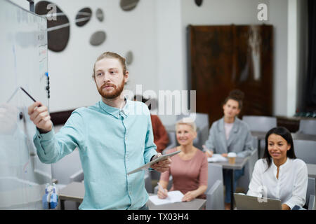 Waist up portrait of contemporary bearded man pointing at whiteboard while presenting training course for students in class, copy space Stock Photo
