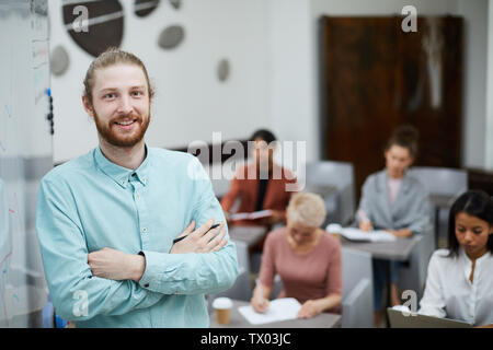 Waist up portrait of contemporary male teacher smiling at camera while standing by whiteboard with group of students in background, copy space Stock Photo