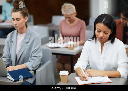 Portrait of mixed-race businesswoman writing notes while sitting at table in class with group of students, copy space Stock Photo