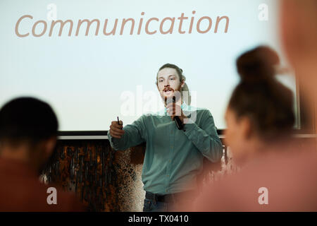 Waist up portrait of contemporary bearded man giving presentation standing by word COMMUNICATION on projector screen and talking to audience during le Stock Photo