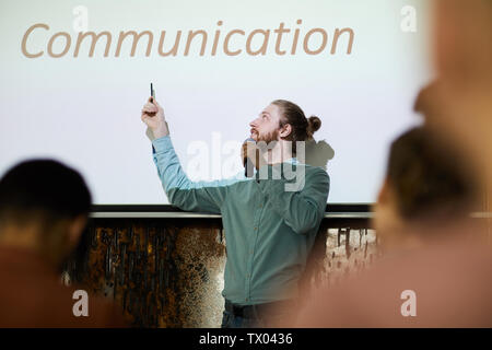 Waist up portrait of contemporary bearded man giving presentation pointing at word COMMUNICATION on projector screen during lecture, copy space Stock Photo