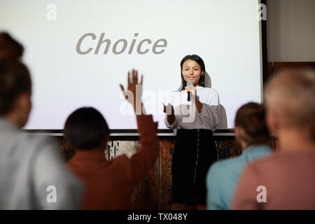 Portrait of successful businesswoman giving speech at conference, with people in audience raising hands, copy space Stock Photo