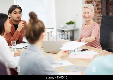 Portrait of mature businesswoman handing papers to colleagues while discussing strategy during meeting in office, copy space Stock Photo