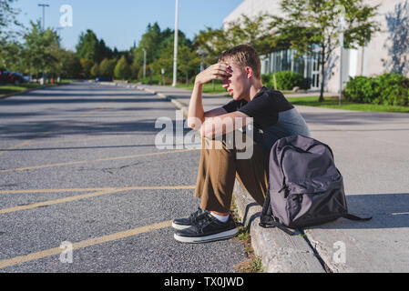 Upset teenager resting his head on his hand as he sits on the curb in front of his school. Stock Photo