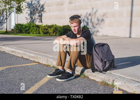 Sad teenage boy sitting on curb near school while listening to music on his headphones. Stock Photo