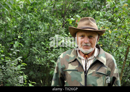 Middle aged traveler. Portrait of a handsome adult man with a gray beard and hat in camouflage clothing against the background of the forest Stock Photo