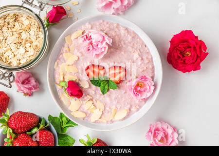 overnight oats or oatmeal porridge with fresh strawberries, almonds and mint in a bowl with rose flowers on white marble table. healthy breakfast. top Stock Photo