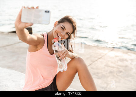 Cheerful young sportswoman resting after workout at the beach, taking a selfie, drinking water Stock Photo