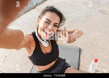 Cheerful young sportswoman resting after workout at the beach, taking a selfie, drinking water, sitting on a fitness mat Stock Photo