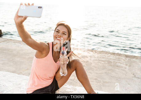 Cheerful young sportswoman resting after workout at the beach, taking a selfie, drinking water Stock Photo