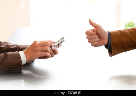 Hands from two businessmen in conversation by a desk. One texting on a mobile phone. The other gives ok sign thumbs up. Negotiating business or a job Stock Photo