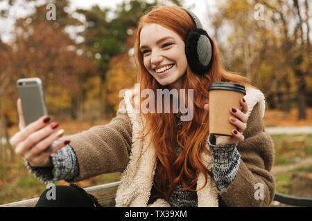 Lovely redheaded young girl listening to music with headpones while sitting on a bench, taking a selfie Stock Photo