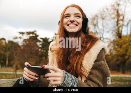 Lovely redheaded young girl listening to music with headphones while sitting on a bench, using mobile phone Stock Photo