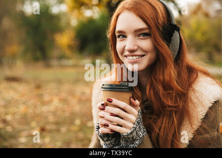 Lovely redheaded young girl listening to music with headpones while sitting on a bench, drinking coffee Stock Photo