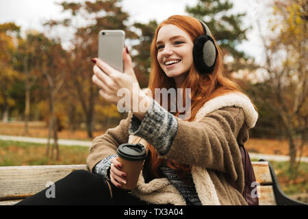Lovely redheaded young girl listening to music with headpones while sitting on a bench, taking a selfie Stock Photo