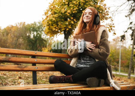 Lovely redheaded young girl listening to music with headpones while sitting on a bench, reading a book Stock Photo