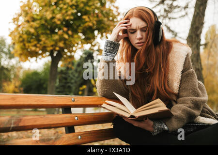 Lovely redheaded young girl listening to music with headpones while sitting on a bench, reading a book Stock Photo