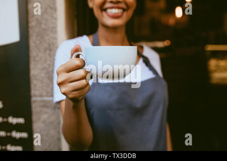 Close up of female barista offering a cup of coffee while standing at the coffeeshop doorway. Focus on cup of coffee in hand of a female cafe owner. Stock Photo