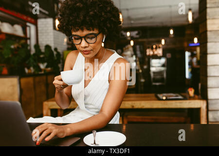 Young woman sitting in cafe working on laptop and drinking coffee. African woman having coffee while working on laptop computer in coffee shop. Stock Photo