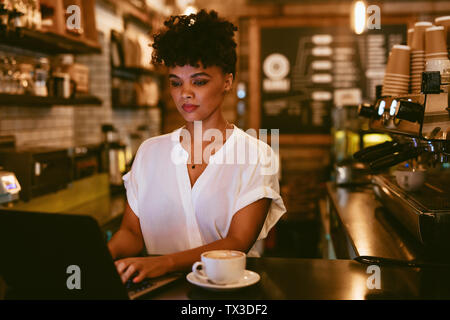 Female cafe owner standing at counter working on laptop with a cup of coffee. Young woman using laptop at her coffee shop.