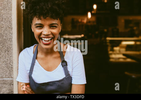 Cafe owner standing in the doorway of her coffee shop. Cheerful young female barista standing with her arms crossed at the door of a restaurant and lo Stock Photo