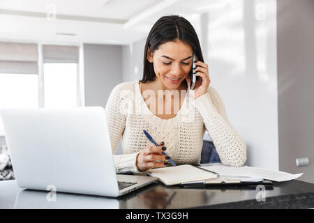 Attractive young woman working with laptop computer and documents while sitting at the kitchen Stock Photo