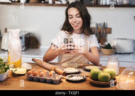 Cheerful young woman wearing apron preparing dough for an apple pie at the kitchen at home, using mobile phone Stock Photo