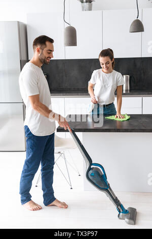 Happy young couple doing general clean at the kitchen, man vacuuming the floor, woman cleaning kitchen desk Stock Photo