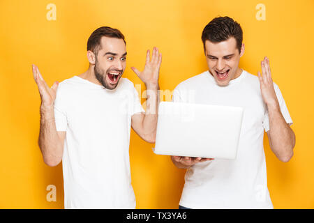 Two cheerful excited men friends wearing blank t-shirts standing isolated over yellow background, using laptop computer, celebrating success Stock Photo