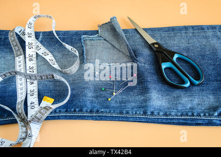 Blue jeans with a large hole on a pant leg below the knee, multi coloured headed sewing pins, white tailor tape with inches and centimeters and scisso Stock Photo