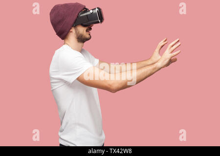 Side view portrait of shocked bearded young man in white shirt and casual hat standing, wearing vr, playing video game and trying to touch something. Stock Photo