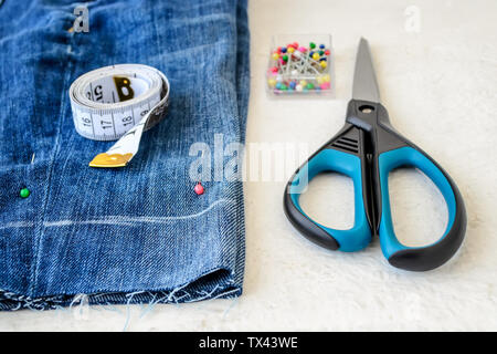 Blue denim shorts, roll of tailor tape with centimeters and inches, multi coloured headed sewing pins in a box and scissors. Making denim shorts. On a Stock Photo