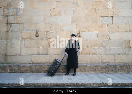 Mature businesswoman wearing black coat and leather cap standing on pavement with rolling suitcase Stock Photo