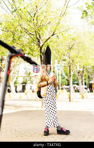 Girl wearing golden sequin jacket, polka dot jumpsuit and black crown drinking soft drink on the street Stock Photo