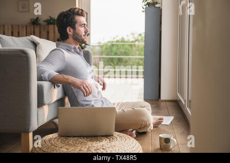 Young man sitting at home on floor, using laptop, looking out of window Stock Photo