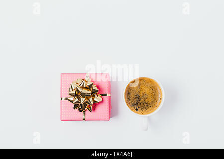 Flat layout of cup of coffee next to pink gift box with golden bow on white background, overhead view. Stock Photo