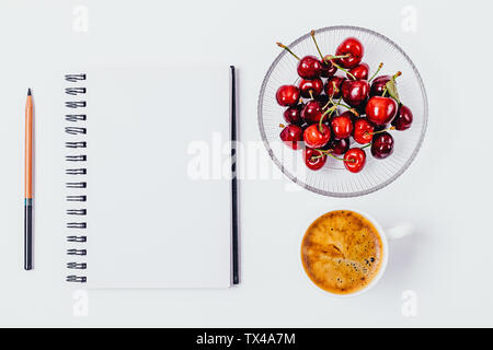 Flat lay composition of blank empty notebook, plate of cherries and cup of coffee on white desktop. Stock Photo