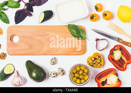 Top view table with wooden board next to ingredients for greek salad, tomatoes, cucumbers, bell pepper, onion, feta cheese and herbs. Stock Photo