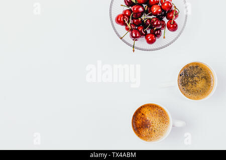 Flat lay composition of red ripe cherries in glass plate next to two cups of freshly brewed coffee on white background with copy space. Stock Photo
