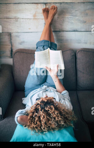 Woman lying on couch at home reading a book Stock Photo