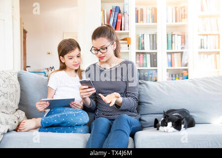 Two sisters sitting on the couch at home using electronic devices Stock Photo