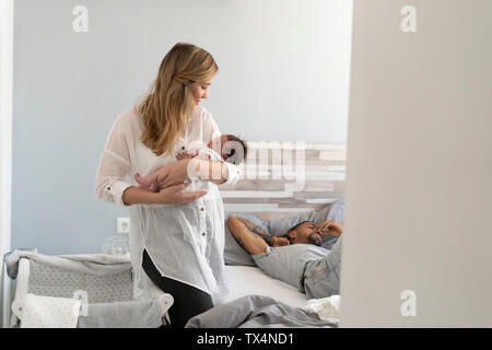Mother holding her newborn baby in bedroom with father sleeping in background