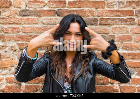 Portrait of young woman wearing black leather jacket and showing rock and roll sign, brick wall