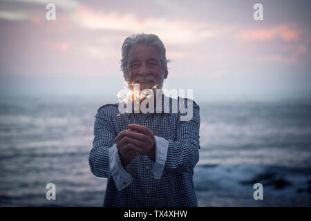 Happy senior man standing in front of the sea by sunset holding sparkler