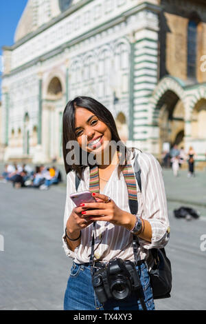 Italy, Florence, portrait of happy young tourist with camera and backpack using smartphone Stock Photo