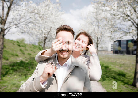 Happy young man holding latchkeys for new house, while woman is covering his eyes Stock Photo