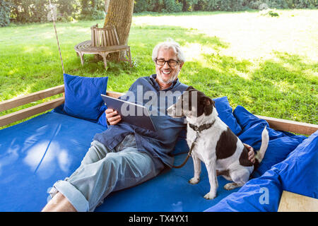 Senior man relaxing on a swing bed in his garden, using digital tablet Stock Photo