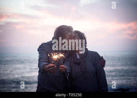 Kissing senior couple standing in front of the sea by sunset holding sparklers Stock Photo