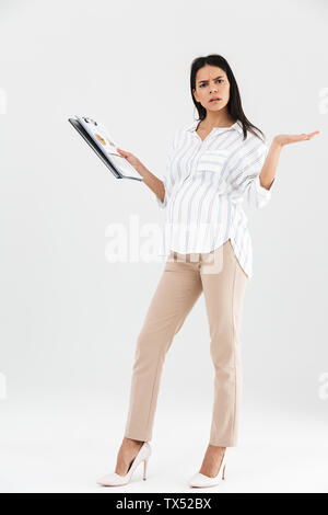 Full length photo of displeased pregnant businesswoman 30s holding clipboard and throwing up hands while standing isolated over white background Stock Photo